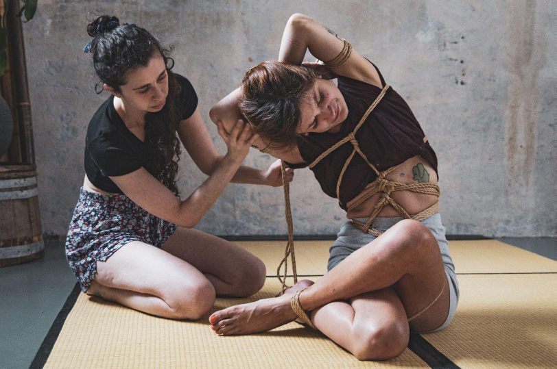 Erato Bänana, a white woman, supports Chocolapain who is bound in a crosslegged position with her arms folded over her head and pulled off balance to her right.