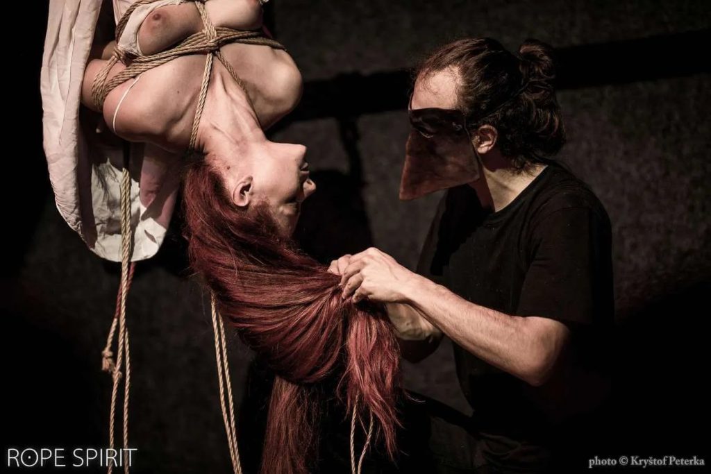 Tenshiko suspended in an inversion has her hair played with by Kirigami who is wearing a mask 